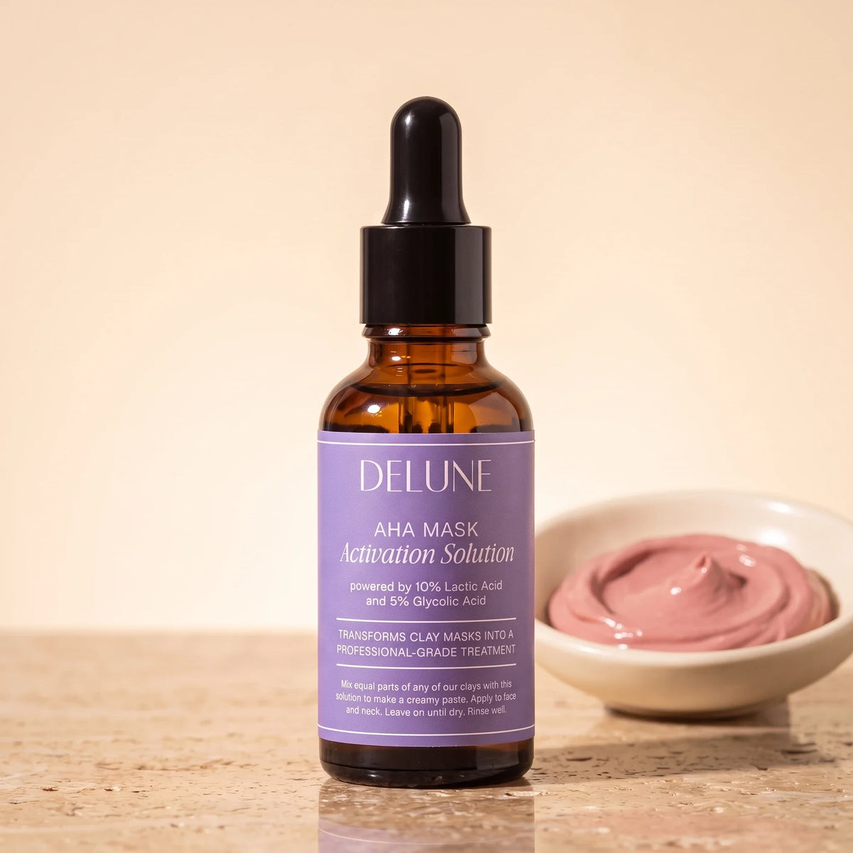 Solución activadora AHA para mascarillas de Delune en frasco gotero de 30 ml sobre travertino, junto a un recipiente con pasta de arcilla rosa