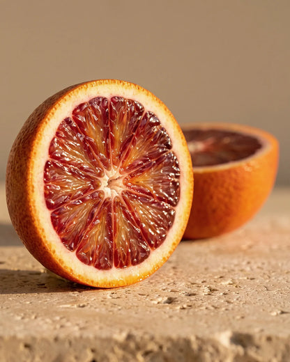 Una naranja sanguina partida a la mitad, con su pulpa rubí intensa, sobre travertino cálido: el aroma cítrico, dulce y afrutado del aceite esencial puro de naranja sanguina de Delune