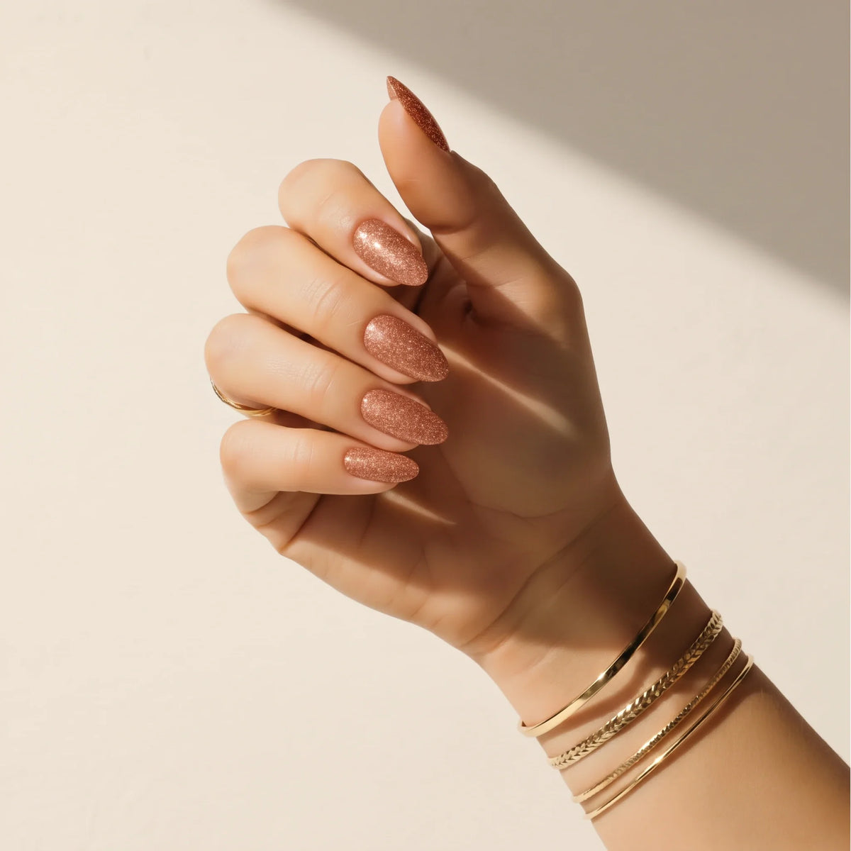 Mano con uñas adhesivas Copper Glitter de Delune y brazaletes dorados sobre una pared de yeso en tono cálido